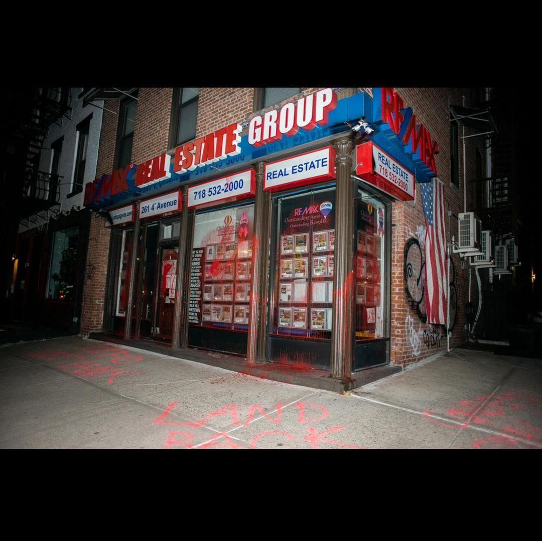 RE/MAX corner storefront splattered with red paint. Graffiti on sidewalk reads "LAND BACK".