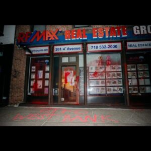 RE/MAX storefront splattered with red paint. Graffiti on sidewalk reads "GTFO WEST BANK".