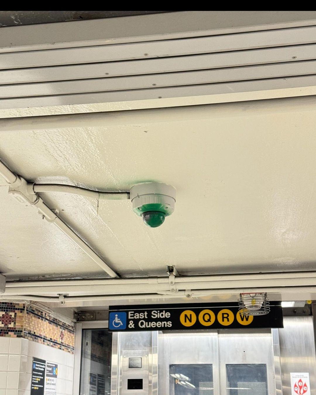 Dome camera on the ceiling of an N/Q/R/W station, covered in green paint