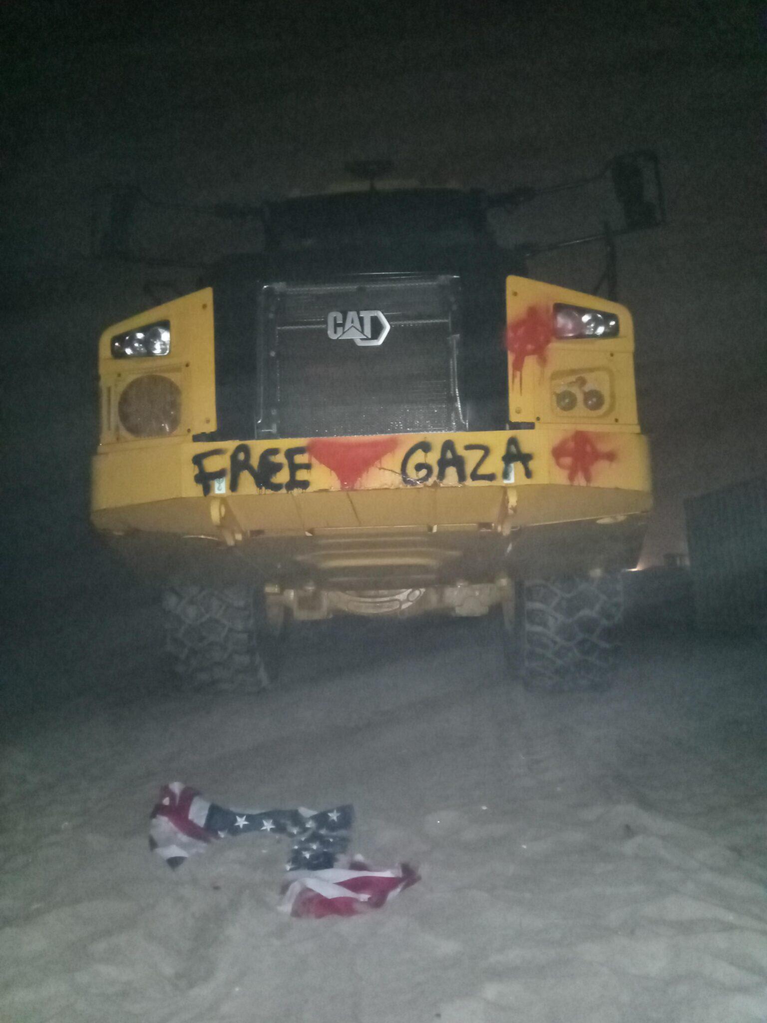A vandalized CAT machine. Graffiti reads "FREE GAZA". There are also circle-A's and a red downward triangle. A torn American flag lies in the foreground.