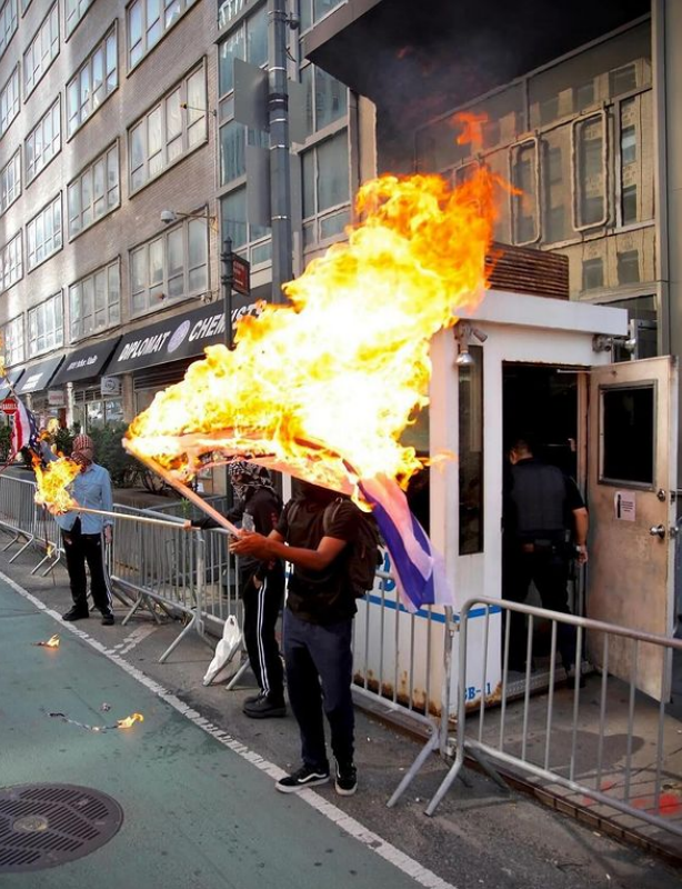 Two people burn the Israeli and US flag next to each other in front an NYPD guard booth.